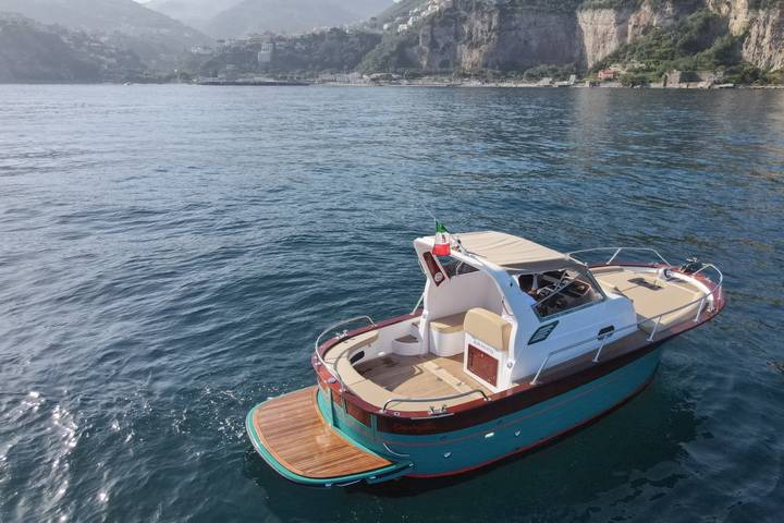 Un gozzo con lo scafo verde acqua, la coperta e la plancetta in teak, all'ancora sull'acqua calma, sullo sfondo le falesie della costa di Sorrento