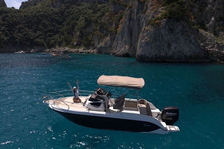 Una lancha motora blanca y negra en agua turquesa bajo los acantilados escarpados y boscosos de Capri, con una pasajera saludando desde la proa